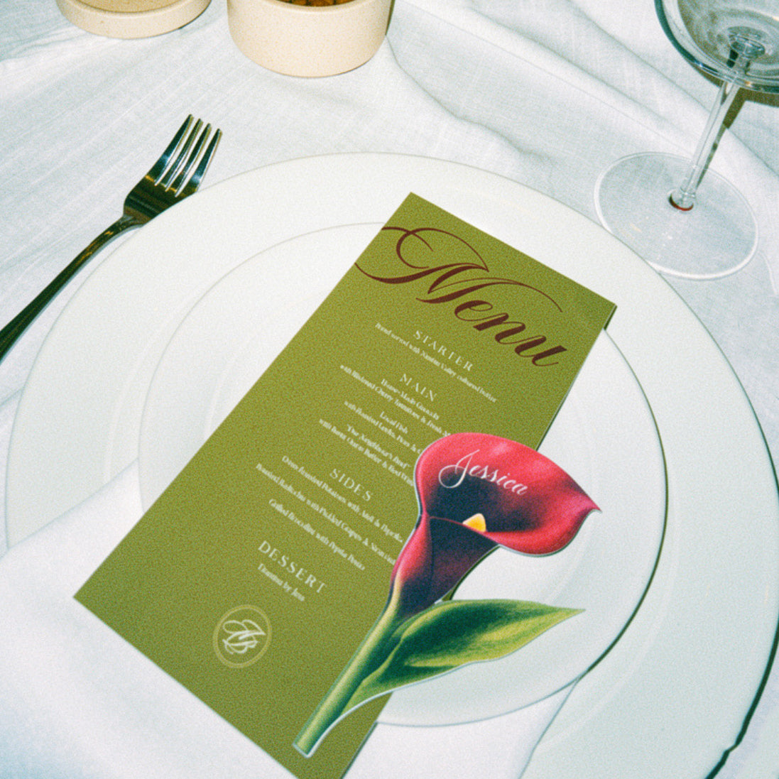 A styled table setting with a white plate holding an olive green menu card tied with a pink ribbon, surrounded by white draped fabric, a cream pleated lamp, and green flowers.