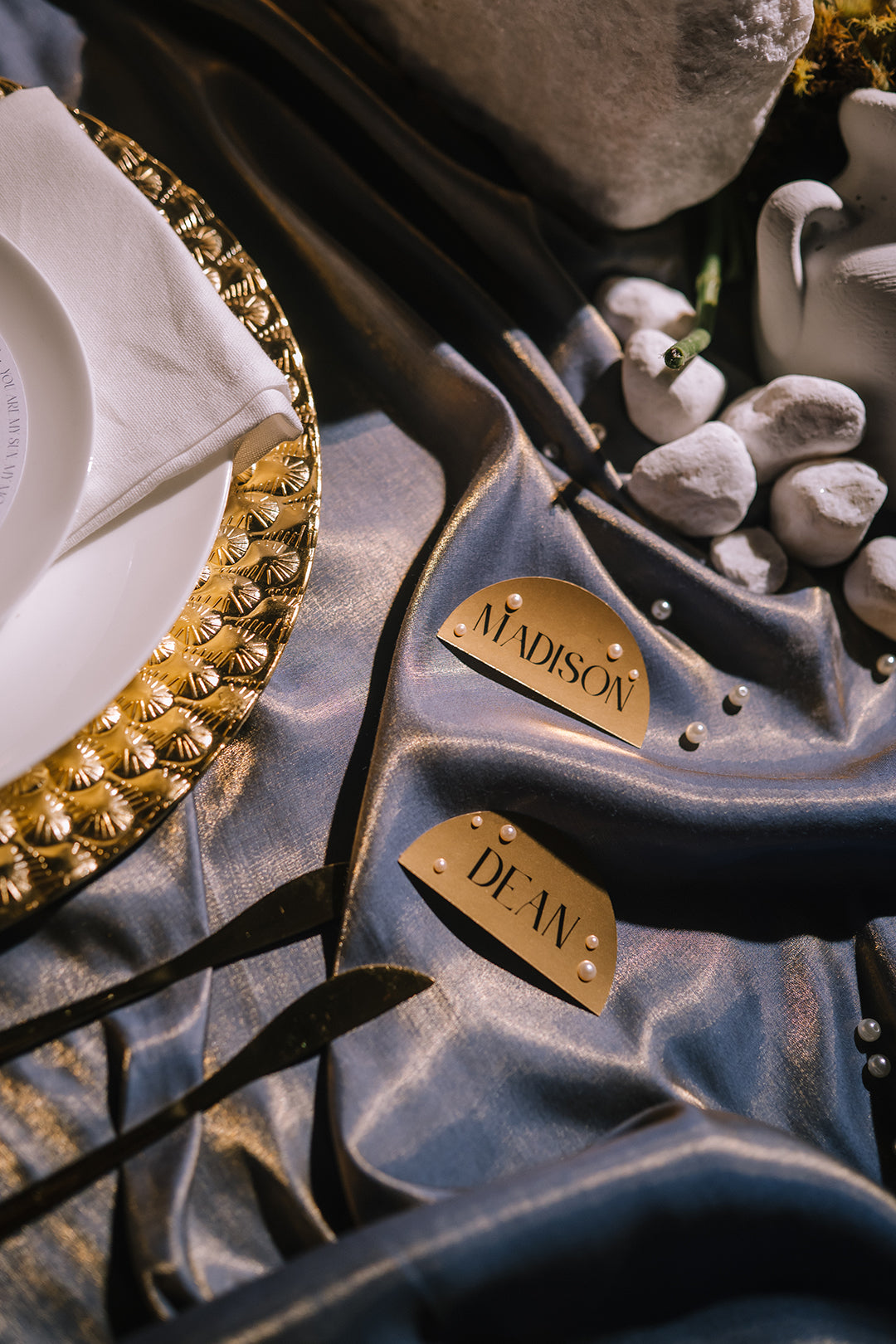 Two personalized place cards reading 'MADISON' and 'DEAN' with small pearl-like accents, styled on a blue satin tablecloth with a gold charger plate, white napkin, and decorative stones.