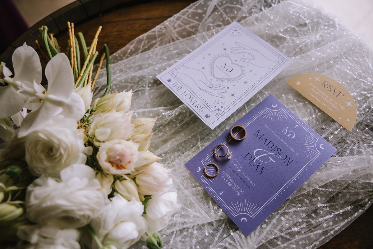 Wedding stationery set arranged on a table with a bouquet of white flowers. The set includes a lavender invitation card, a matching RSVP card in gold, a lavender wedding day card, and two gold wedding rings on a crinkled clear fabric background.