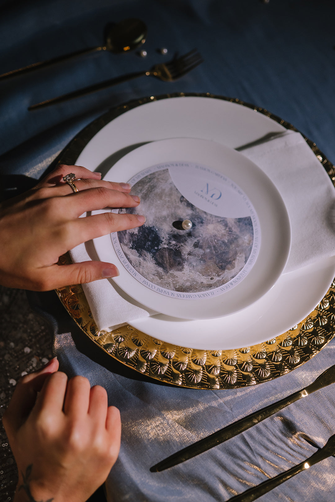 A close-up of a place setting featuring a white dinner plate with a gold scalloped rim, a white napkin, and a round printed insert with a moon illustration and the number 10. A person's hands are visible adjusting the insert, set against a dark blue tablecloth with gold-toned cutlery.