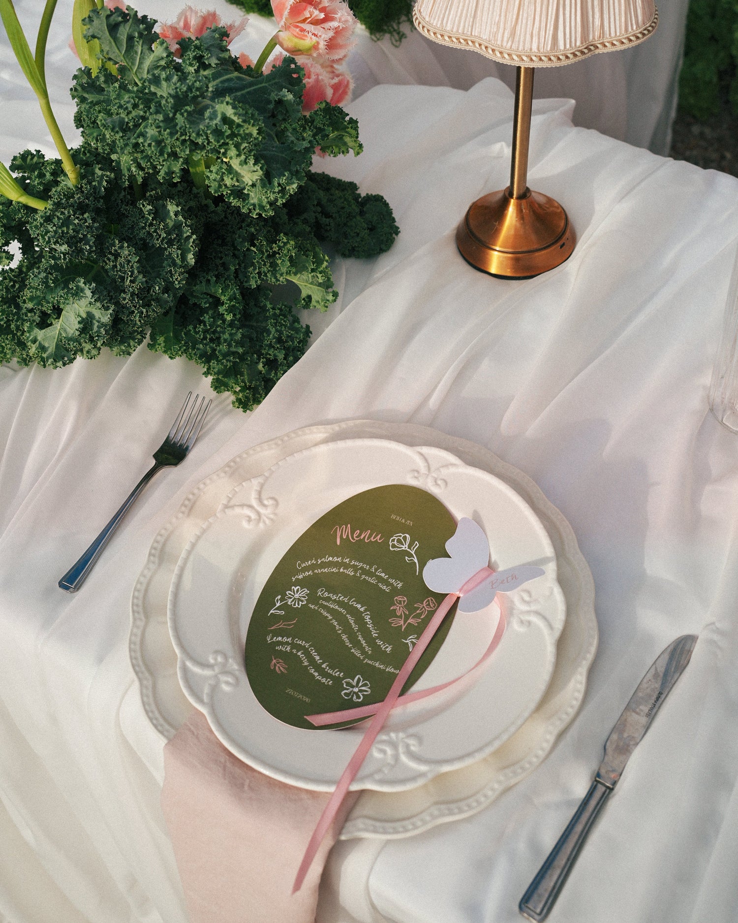 A styled table setting with a white plate holding an olive green menu card tied with a pink ribbon, surrounded by white draped fabric, a cream pleated lamp, and green flowers.